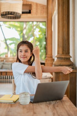 Genç bir kadın, tropikal bir ortamda elinde dizüstü bilgisayar ve defterle bir ofis masasında çalışırken esniyor..