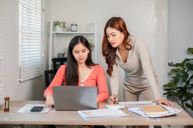 Parlak bir ofiste birlikte çalışan iki kadın dizüstü bilgisayar ve belgeler üzerinde fikir alışverişinde bulunuyor..
