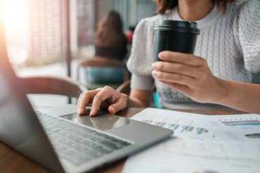 Laptop ve kahveyle birden fazla iş yapan profesyonel bir kadın, çağdaş ofis ortamında finansal belgeleri gözden geçiriyor..