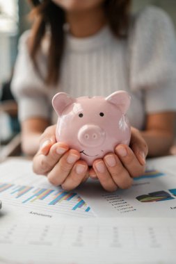Finansal tabloların üzerinde pembe bir kumbarayı elinde tutan bir kadının yakın plan çekimi, birikimleri, bütçeleri ve finansal planlamaları temsil ediyor..