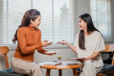 İki kadın, sıcak, güneşli bir odada ahşap mobilyalar ve masada küçük bir bitki eşliğinde canlı bir tartışmanın keyfini çıkarıyorlar..
