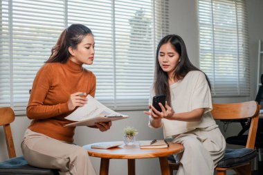 Modern bir ofiste çalışan iki kadın, ahşap bir masada oturarak belgeleri ve akıllı telefonu kullanıyorlar..