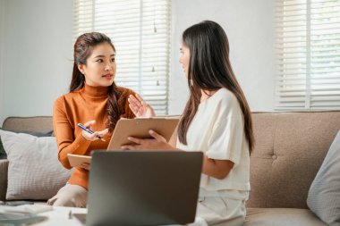 Konforlu bir oturma odasında iş birliği yapmak için dizüstü bilgisayar ve tablet kullanan iki kadın..