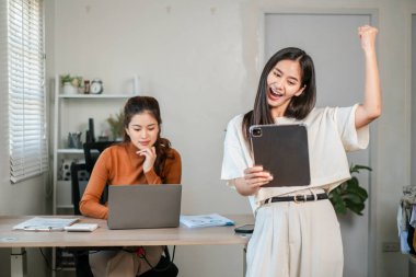 Modern bir ofiste iki kadın, biri tabletle kutlama yapıyor, diğeri dizüstü bilgisayarla çalışıyor..