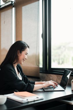Geniş pencereli ve doğal ışığı olan parlak, modern bir ofis ortamında dizüstü bilgisayar kullanan odaklanmış bir kadın..