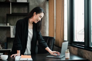 Modern ofis ortamındaki iş kadını, dizüstü bilgisayarına odaklanmış, çağdaş dekor ve doğal ışıkla çevrili..