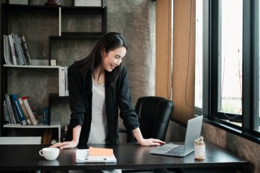 Siyah ceketli bir iş kadını, şık bir ofis ortamında dizüstü bilgisayara gülümsüyor..