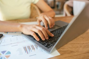 Ahşap bir masanın üzerinde finansal belgelerle bilgisayarda yazı yazan ellerin yakın çekimi, verimli bir çalışma ortamı sergiliyor..