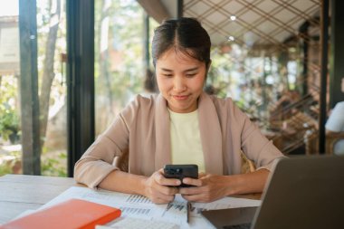 Modern ofis ortamında genç bir kadın, akıllı telefonuna odaklanmış, bir dizüstü bilgisayar ve belgelerle çevrili..