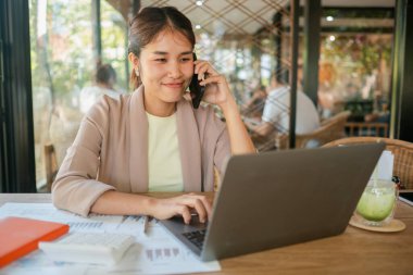 Genç bir kadın bir kafede birden fazla iş yapıyor. Laptop ve akıllı telefon kullanıyor. Etrafı dokümanlarla çevrili..