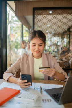 Akıllı telefon ve kredi kartıyla evdeki ofis masasında bilgisayarlı ve belgeli alışveriş yapan gülümseyen bir kadın..