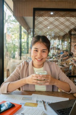 Modern bir kafe ortamında elinde kahve fincanı tutan gülümseyen bir kadın. Etrafı dokümanlar ve dizüstü bilgisayarlarla çevrili..