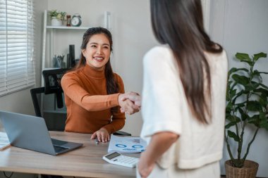 Modern ofis ortamındaki iki kadın profesyonel bir tokalaşma içinde, başarılı iş ortaklığını simgeliyor..