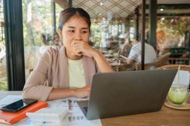 Belgeler ve ferahlatıcı bir içecek ile çevrili, parlak bir kafede dizüstü bilgisayarına derinden odaklanmış bir kadın..