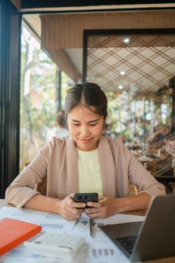 Bir kafede genç bir kadın, akıllı telefonuna odaklanmış, bir laptop ve belgelerle çevrili, modern bir çalışma ortamı sergiliyor..