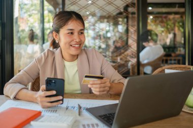Akıllı telefon ve kredi kartı kullanan gülümseyen bir kadın bilgisayarlı ofis masasında online alışveriş yapıyor..