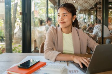 Modern bir kafede çalışan genç bir kadın. Bilgisayarında iş belgeleri ve akıllı bir telefonla çalışıyor. Etrafı ışık saçıyor..
