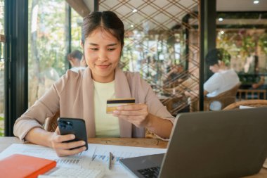 Genç bir kadın bilgisayarlı ve belgeli bir kafede otururken akıllı telefonu ve kredi kartını online alışveriş için kullanıyor..