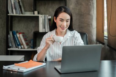 Çizgili tişörtlü, elinde kahve fincanı olan gülümseyen bir kadın, çağdaş ofis ortamında bir masada dizüstü bilgisayar üzerinde çalışıyor..