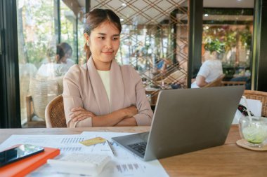 Bir kafede çalışan genç bir kadın. Etrafı dokümanlar ve hesap makinesiyle çevrili. Modern bir çalışma ortamı sergiliyor..