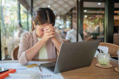 Bir kafede genç bir kadın, iş konusunda stresli, etrafı dizüstü bilgisayar ve belgelerle çevrili, çözüm arayan.