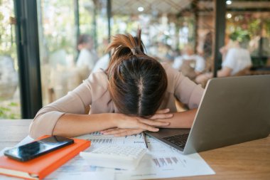 Yorgun bir kadın, başını bir masanın üzerinde dinlendiriyor. Etrafı dizüstü bilgisayarla, belgelerle ve güneşli bir ofiste akıllı bir telefonla çevrili..