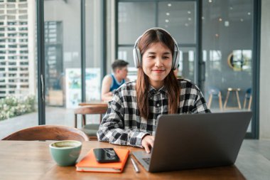 Ekose gömlekli genç bir kadın modern bir kafede dizüstü bilgisayar kullanıyor, gülümsüyor ve uzaktan çalışıyor..
