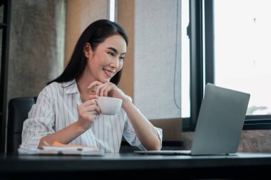 İş kadını elinde kahve fincanı tutarken gülümsüyor ve parlak, modern bir ofiste dizüstü bilgisayarla çalışıyor..