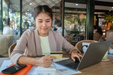 Evraklar ve teknolojik cihazlarla çevrili rahat bir kafede online alışveriş yapmak için dizüstü bilgisayar ve kredi kartı kullanan bir kadın..