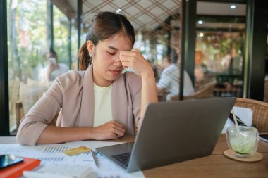 Bir kafede genç bir kadın, bir dizüstü bilgisayar üzerinde çalışırken stresli, belgelerle çevrili, ve ferahlatıcı bir içecek ile.