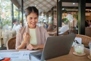 Kafede gülümseyen bir kadın, heyecanlı bir şekilde dizüstü bilgisayarına bakıyor, etrafı kağıtlarla çevrili ve ferahlatıcı bir içecekle dolu..