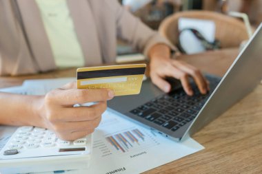 Kredi kartını tutan, online alışveriş için dizüstü bilgisayar kullanan, mali belgeleri ve hesap makinesiyle ahşap bir masada duran bir elin yakın çekimi..