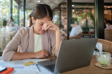 Belgeler ve kredi kartıyla çevrili modern bir kafede dizüstü bilgisayarla çalışan genç bir kadın stresli görünüyor..