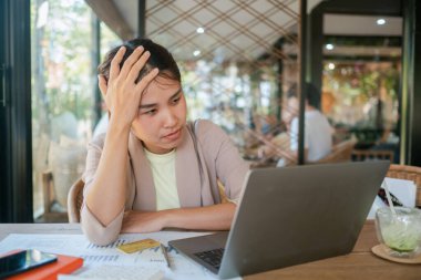 Modern bir kafe ortamında dizüstü bilgisayarla çalışırken stresli görünen genç bir kadın, etrafı gazeteler ve bir fincan kahveyle çevrili..