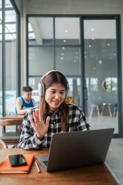 Bir kafede dizüstü bilgisayar ve kulaklık kullanan genç bir kadın, video görüşmesi sırasında el sallıyor. Uzak çalışma ya da çevrimiçi haberleşme ile meşgul.