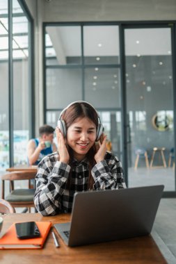Kulaklık takan, çağdaş bir kafe ortamında dizüstü bilgisayar kullanan, müzikten ve işten zevk alan gülümseyen bir kadın..