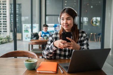 Modern bir kafede akıllı telefon ve dizüstü bilgisayar kullanan kulaklıklı bir kadın. Rahat ve verimli bir atmosfer..