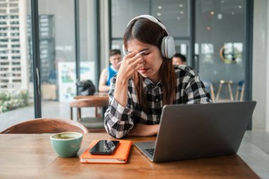 Bir kafede genç bir kadın, dizüstü bilgisayarda çalışırken stresli kulaklık ve ekose gömlek takıyor..