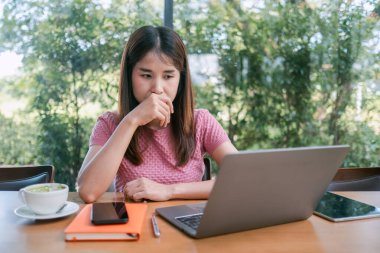 Yeşilliklerle çevrili bir kafede dizüstü bilgisayarda çalışan akıllı bir kadın. Masasında akıllı telefon, tablet ve not defteri var..