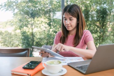 Bir kafede, tablet kullanan genç bir kadın. Masasında laptopu ve kahveyle, yeşillikle çevrili..