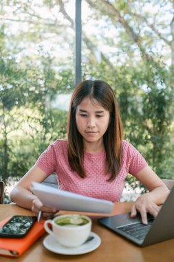 Yeşillik, belgeler ve kahveyle çevrili bir kafede dizüstü bilgisayarıyla çalışan odaklanmış bir kadın..