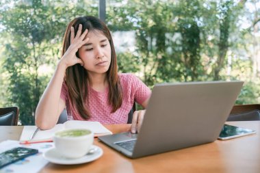 Bir kafede dizüstü bilgisayarı ve yeşil çayıyla çalışan genç bir kadın. Etrafı büyük pencerelerden yeşilliklerle çevrili..