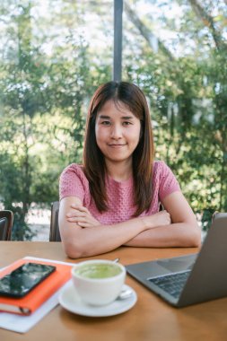 Genç bir kadın, elinde laptop ve matcha latte ile bir kafe masasında oturuyor, doğal ışık ve yeşillikle huzurlu bir çalışma ortamının tadını çıkarıyor..