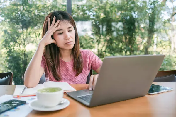 Bir kafede dizüstü bilgisayarı ve yeşil çayıyla çalışan genç bir kadın. Etrafı büyük pencerelerden yeşilliklerle çevrili..