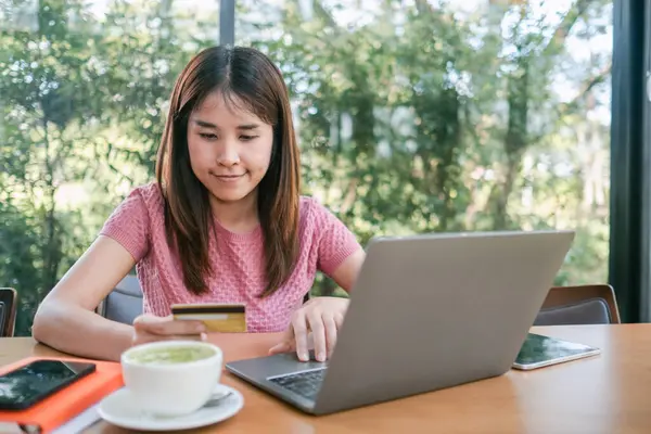 Çevresi yeşilliklerle çevrili bir kafede dizüstü bilgisayar ve kredi kartı kullanan bir kadın internetten alışveriş yapmaktan hoşlanıyor..