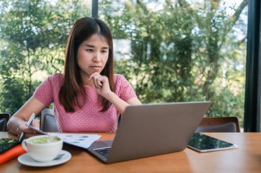 Yeşilliklerle çevrili bir kafede dizüstü bilgisayara odaklanan genç bir kadın. Masasında kahve ve belgeler var..