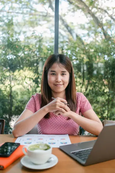 Bir kafede laptopla çalışan genç bir kadın, kahvesinin tadını çıkarıyor, arka planda yeşillik ve doğal ışık var..