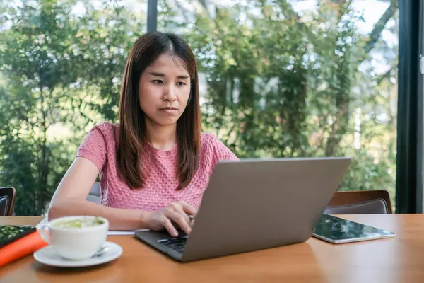 Yeşillik ve doğal ışıkla çevrili bir kafede dizüstü bilgisayarına odaklanan genç bir kadın..