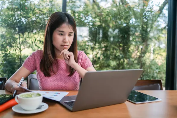Yeşilliklerle çevrili bir kafede dizüstü bilgisayara odaklanan genç bir kadın. Masasında kahve ve belgeler var..