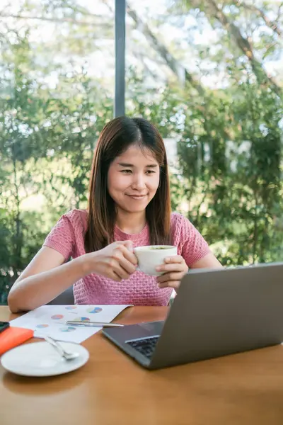 Elinde kahve fincanı olan gülümseyen bir kadın, dışarıda yeşil bir kafede dizüstü bilgisayarla çalışıyor..